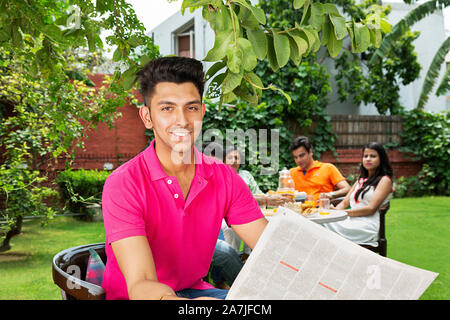 Smiling Adult man Sitting-in-Chair And Reading Newspaper with family in background at courtyard of their home Stock Photo