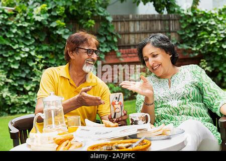 happy senior Couple Husband and wife enjoying Breakfast and Using Cell-phone and talking in-courtyard near their houseÂ Stock Photo
