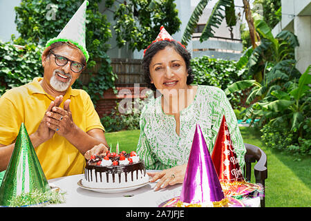 Happy Old couple celebrating birthday and cutting birthday cake at-courtyard of-their home Stock Photo