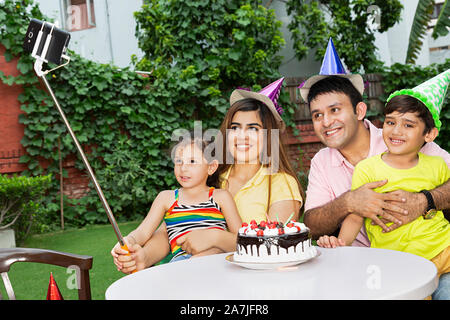 Happy-family Parents And Two-Children taking Selfie picture with smartphone on selfie stick during birthday celebration in-Courtyard Of-Their Home Stock Photo