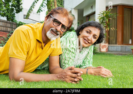 Portrait Of Smiling Senior Couple lying-on-grass and relaxing at-courtyard of-their home Stock Photo