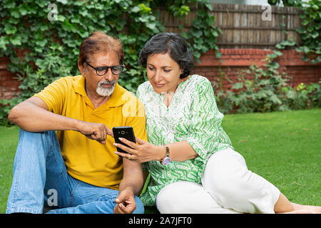 Happy Elderly Couple Relaxing Sitting on-grass Using Cell Phone In-garden Stock Photo