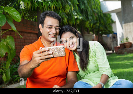 Happy Mid-Adult Couple taking a selfie Photo By Cell Phone And sitting-on-grass at-courtyard of-their home Stock Photo
