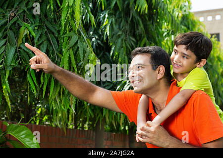 Happy Indian father holds his Kid son piggyback ride on his shoulders points at something in-Outdoor garden Stock Photo