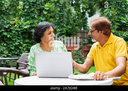 Happy indian Senior Couple Male And Female Using Laptop And With Newspaper in the courtyard of their house On Summer Morning Stock Photo