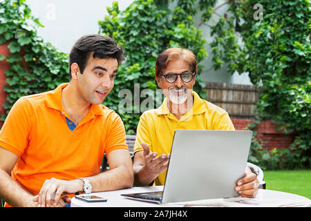 Senior Father And Adult Son Sitting on-table Using laptop Together in-courtyard of their house Stock Photo