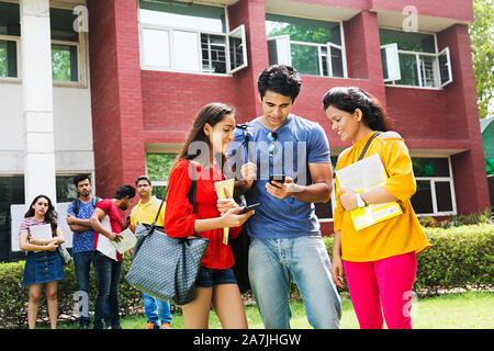 Three Young Male And Female College Students Friends Together Reading Text-message Cell-Phone In-Outside Campus Building Stock Photo