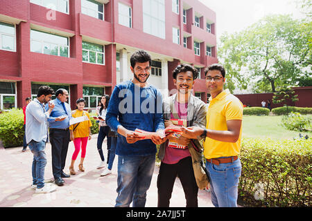 Three College Boys Students Friends Reading Book Studying Together With People-In-Background In-Outside Campus Building Stock Photo