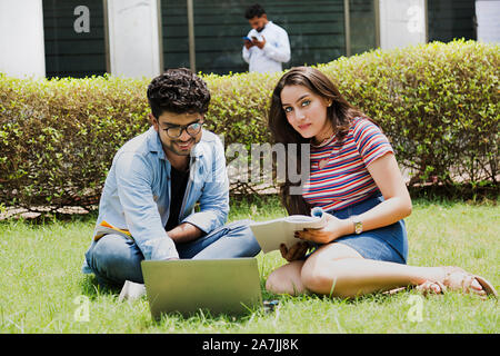 Two Young College Couple Students Studying Book With Laptop Education Learning Sitting-on-Grass At-Campus Stock Photo