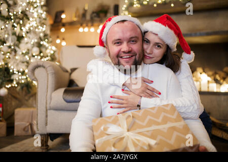 Picture of man and woman in Santa's caps sitting on floor with gifts in room with New Year's interior Stock Photo