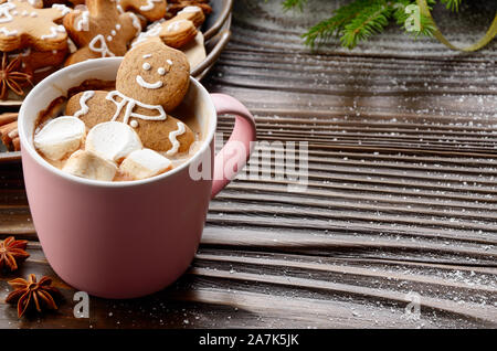 Pink mug with hot chocolate marshmallows and gingerbread man on background of spruce branch and tray with cookies Stock Photo