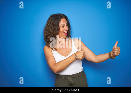 Middle age senior woman with curly hair standing over blue isolated background Looking proud, smiling doing thumbs up gesture to the side Stock Photo