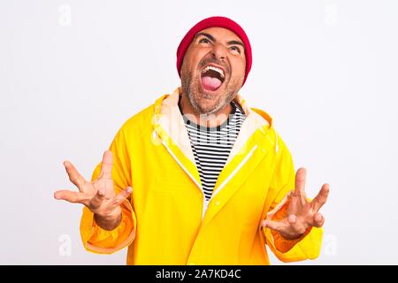 Middle age man wearing rain coat and woolen hat standing over isolated white background crazy and mad shouting and yelling with aggressive expression Stock Photo