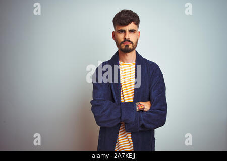 Young man with tattoo wearing bathrobe standing over isolated white background skeptic and nervous, disapproving expression on face with crossed arms. Stock Photo