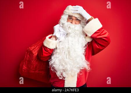 Middle age man wearing Santa costume holding sack with gifts over isolated red background stressed with hand on head, shocked with shame and surprise Stock Photo