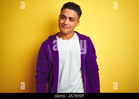 Young brazilian man wearing purple sweatshirt standing over isolated yellow background smiling looking to the side and staring away thinking. Stock Photo