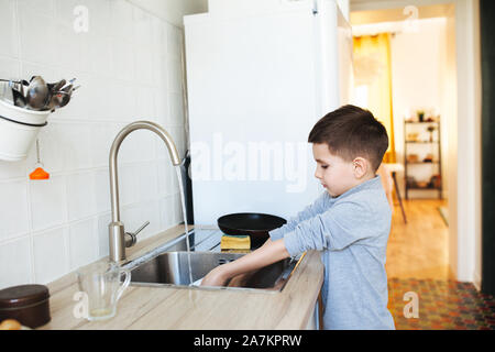 Little cute boy washing dishes in kitchen interior. Child with helping his parents with housework. Stock Photo
