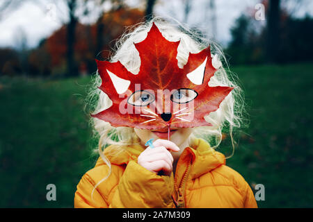 Portrait of cute child kid with blue eyes holding autumn fall red maple leaf painted mask in front face. Girl playing pretending to be fox cat. Fairy Stock Photo
