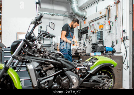 Handyman in overalls working at the workplace with lots of insrtuments at the workshop with motorcycle on the foreground Stock Photo