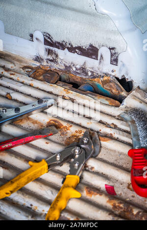 Rusty hole in vehicle floor with tools in the foreground, tin scissors, wire brush and chisel Stock Photo