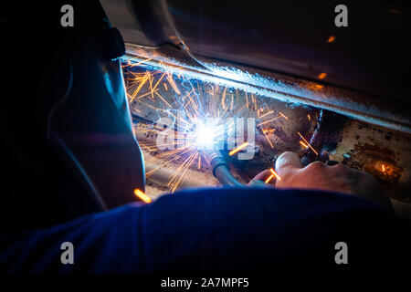 Man with a helmet and no gloves welds in the metal of a car and repairs a rusty hole Stock Photo