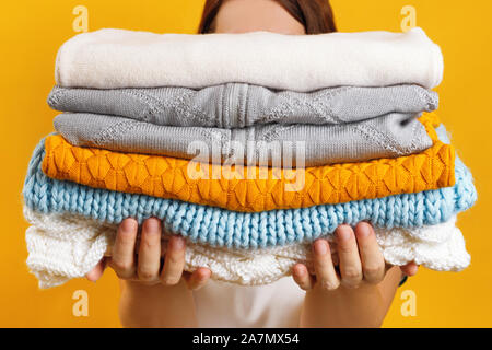 A woman holds a stack of warm knitted clothes on a background of a yellow wall. Stock Photo