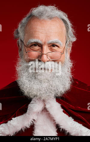 Head and shoulders portrait of classic bearded Santa looking at camera and smiling while posing against red background in studio, copy space Stock Photo