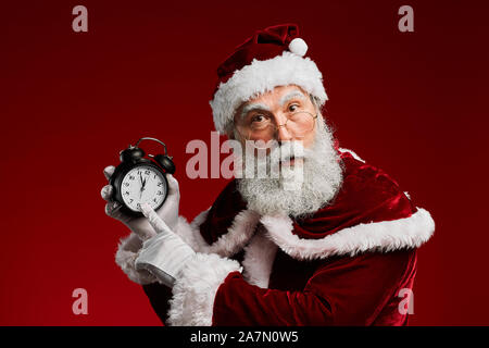 Waist up portrait of classic Santa Claus pointing at clock while posing against red background in studio, copy space Stock Photo