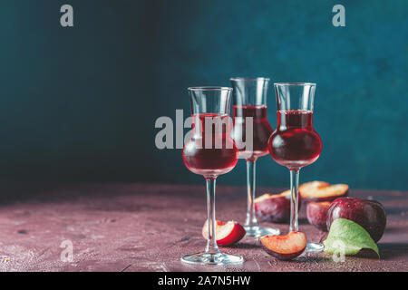 Plums strong alcoholic drink in grappas wineglass with dew. Hard liquor, slivovica, plum brandy or plum vodka with ripe plums on dark blue and  claret Stock Photo