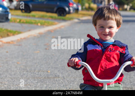A young toddler, a 2-year-old, rides a tricycle down a suburban street. The little boy smiles as he goes, wearing a light jacket for fall or spring we
