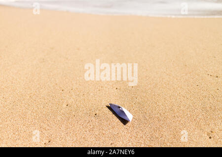 Boat origami stranded on a beach, with foamy wave on the background Stock Photo