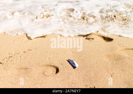 Boat origami stranded on a beach, with foamy wave on the background Stock Photo