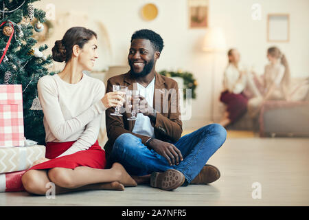 Full length portrait of modern adult couple sitting by Christmas tree and clinking champagne glasses at home, copy space Stock Photo