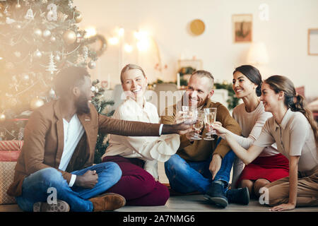 Full length portrait of cheerful friends clinking champagne glasses sitting by Christmas tree during party, copy space Stock Photo