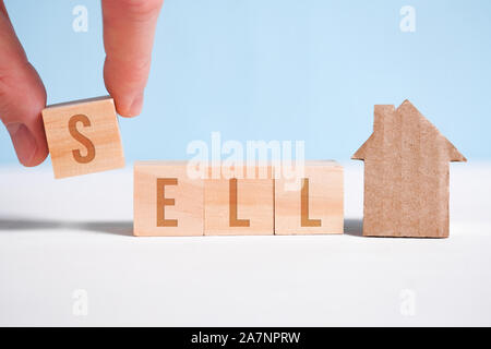 Abstract house made of cardboard hand holds with wooden cubes with the word sell. Home sale concept. Close up. Stock Photo