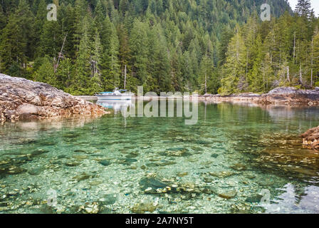 A power boat, anchored and stern tied in a quiet, forested coastal inlet in British Columbia, is visible over a half-submerged reef in the foreground. Stock Photo