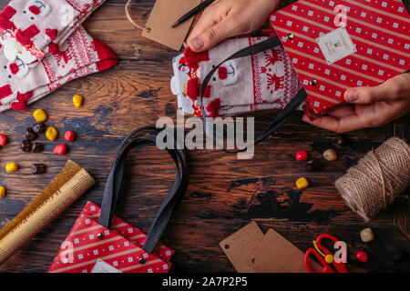 Christmas background. Top view of woman hands wrap New Year present and tie bow. Packed gifts and scrolls, spruce branches and tools on shabby wooden Stock Photo