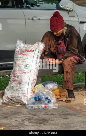 Da Nang, Vietnam - March 10, 2019: Homeless woman sits on bench along street with bags of recycled trash. Stock Photo