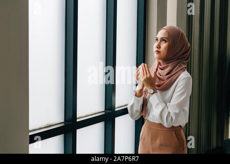 Southeast Asian Islamic female in hijab clasping hands and looking up while standing near window during prayer Stock Photo