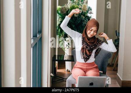 Happy Asian woman in hijab raising arms and stretching body while working with laptop in cozy cafe Stock Photo