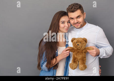 Relationship Concept. Young couple studio standing isolated on grey with teddy bear posing to camera cute Stock Photo