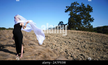 very pregnant woman wearing long stretchy dress, a long white scarf around face blowing in the wind, standing on dirt field with large fallen tree Stock Photo