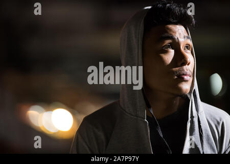 Face of young Asian man with hoodie thinking in the city streets at night Stock Photo