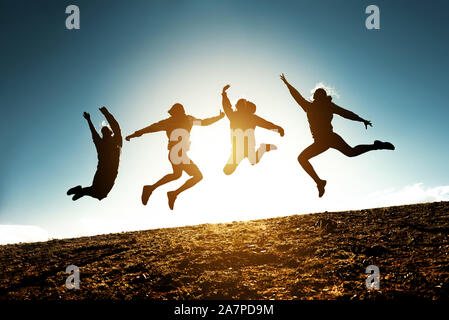 Four silhouettes of friends are jumping together against blue sky and sun Stock Photo