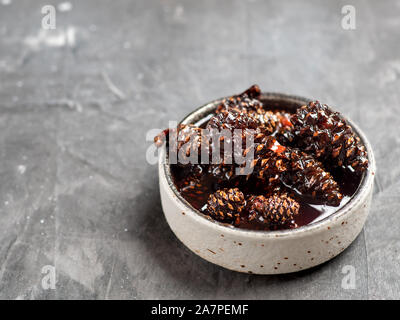 Delicious jam with baby pine cones in small bowl. Traditional Siberian dessert - young pine cones jam on gray textured background. Stock Photo
