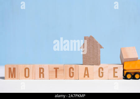 Cardboard cutout house on cubes with inscription mortgage and toy truck on blue background. Concept of construction loan, hypothec, debt, credit. Stock Photo