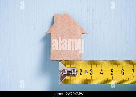 Cardboard cutout house on metal measuring tape on wooden blue background. Concept of surveying, budgeting, repairing. Stock Photo