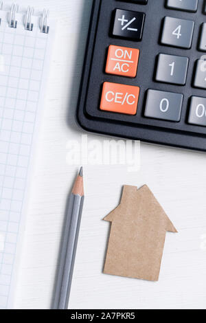Cardboard cutout house, pencil, calculator and blank sheet of spiral notebook on white background. Vertical. Concept of construction, budgeting, repai Stock Photo