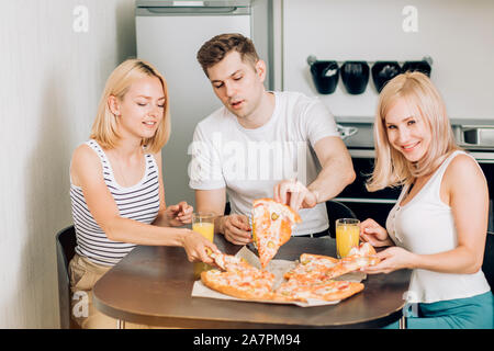 Friendship, people and food concept. Three caucasian happy students eating pizza in kitchen.They are having party at home, laughing and having fun, dr Stock Photo