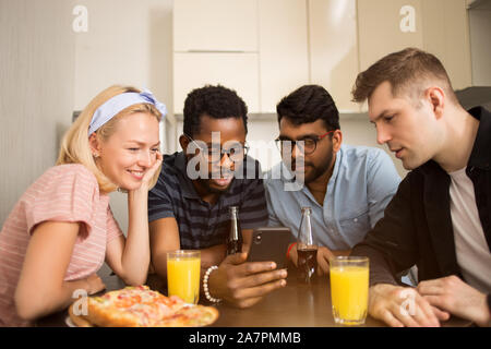 Four friends sitting at table, eating pizza, drinking soft beverages, reading text message on smartphone, testing new phone application together. Mult Stock Photo